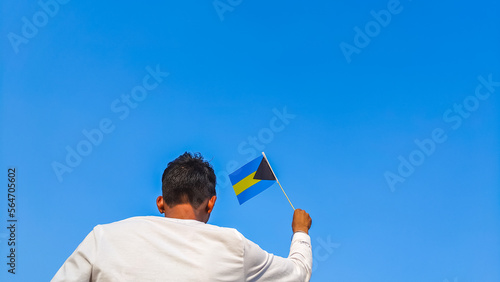 Boy holding Bahamas flag against clear blue sky. Man hand waving Bahamian flag view from back, copy space