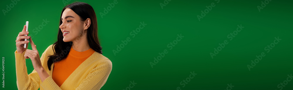 pleased woman in orange turtleneck using smartphone isolated on green, banner. pleased woman in orange turtleneck using smartphone isolated on green, banner.