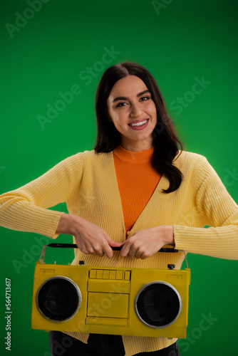 brunette woman in yellow jumper and orange turtleneck holding boombox and looking at camera isolated on green.