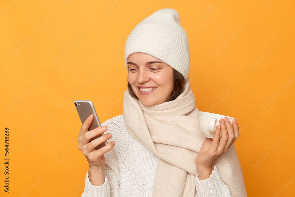Image of attractive caucasian joyful woman wearing sweater, hat and scarf, holding cup of coffee in hands and using cell phone, reading good news in internet, posing isolated over yellow background. Image of attractive caucasian joyful woman wearing sweater, hat and scarf, holding cup of coffee in hands and using cell phone, reading good news in internet, posing isolated over yellow background.