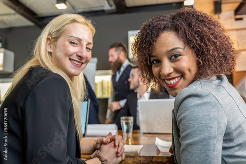 Young black south american woman looking at camera with her blonde caucasian colleague while working in modern office - Break from work and coworking concept © Davide Zanin