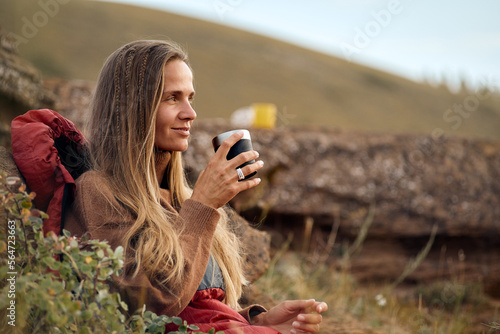 Charming woman sit leaning on big stone relaxing, drinking tea in nature alone, enjoying landscape, dressed in casual warm sweater, looking at side in contemplation. travel, people lifestyle