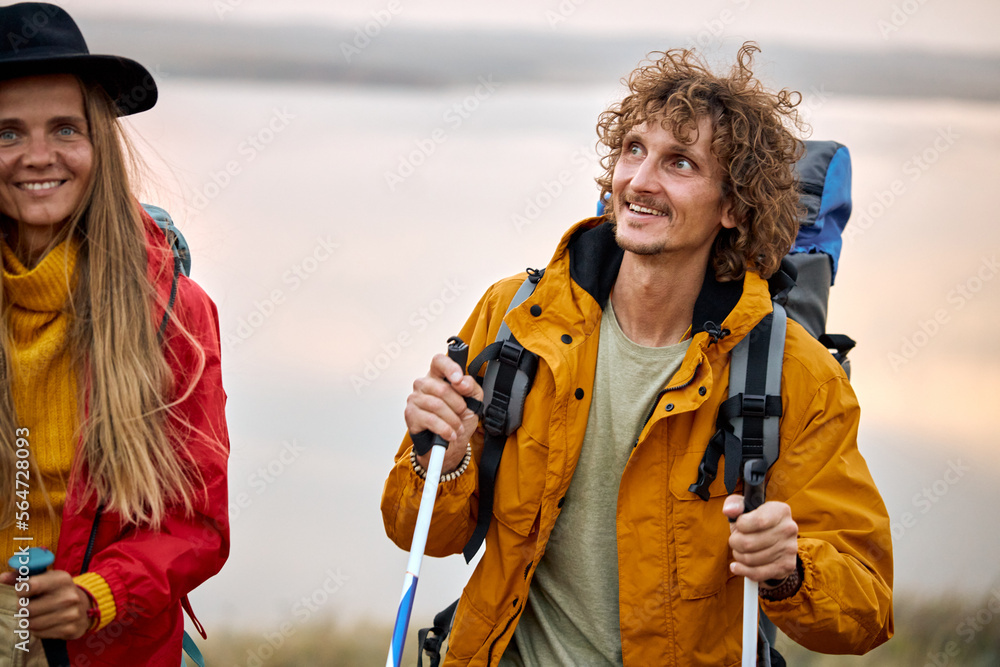 happy young couple walking in the field while hiking with backpacks and trekking sticks. Concept of active lifestyle, sport and travel. caucasian enthusiastic man and woman in sportive clothes.