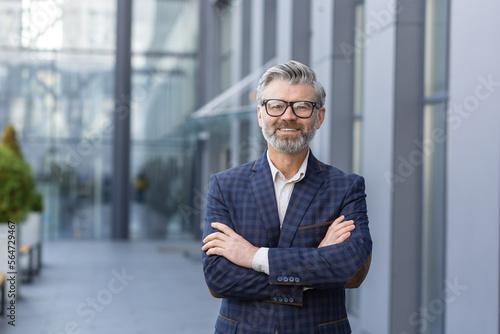 Billede på lærred Portrait of mature gray-haired businessman, senior boss smiling and looking at camera with arms crossed, gray-haired man outside office building, successful investor and owner in business suit