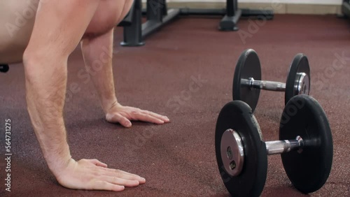 Athletic man is doing push-ups on floor in gym near the dumbbells, side view. Sportsman with muscular body training press ups. Sport, training, bodybuilding, healthy lifestyle, exercising concept. 