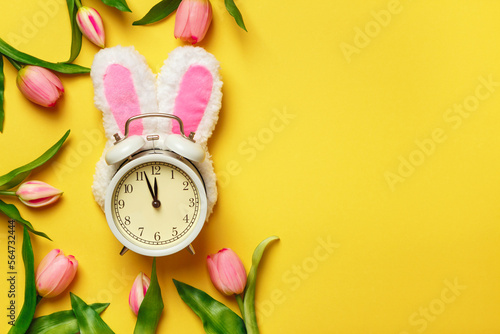 Alarm clock in rabbit ears surrounded by tulips. Easter spring change of seasons.