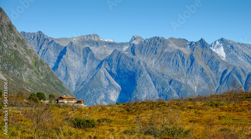 A solitary cabin nestled in a valley, framed by rugged mountains and golden foliage, basking in daylight.