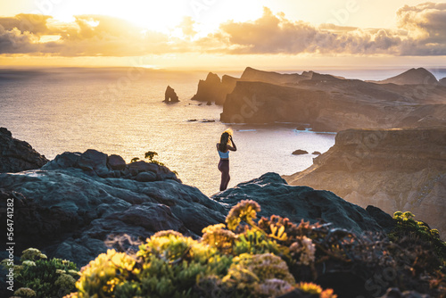 Woman enjoys panoramic view from view point on a steep cliff over the seascape and along the rugged foothills of Madeira coast at sunrise. Ponta do Bode, Madeira Island, Portugal, Europe.
