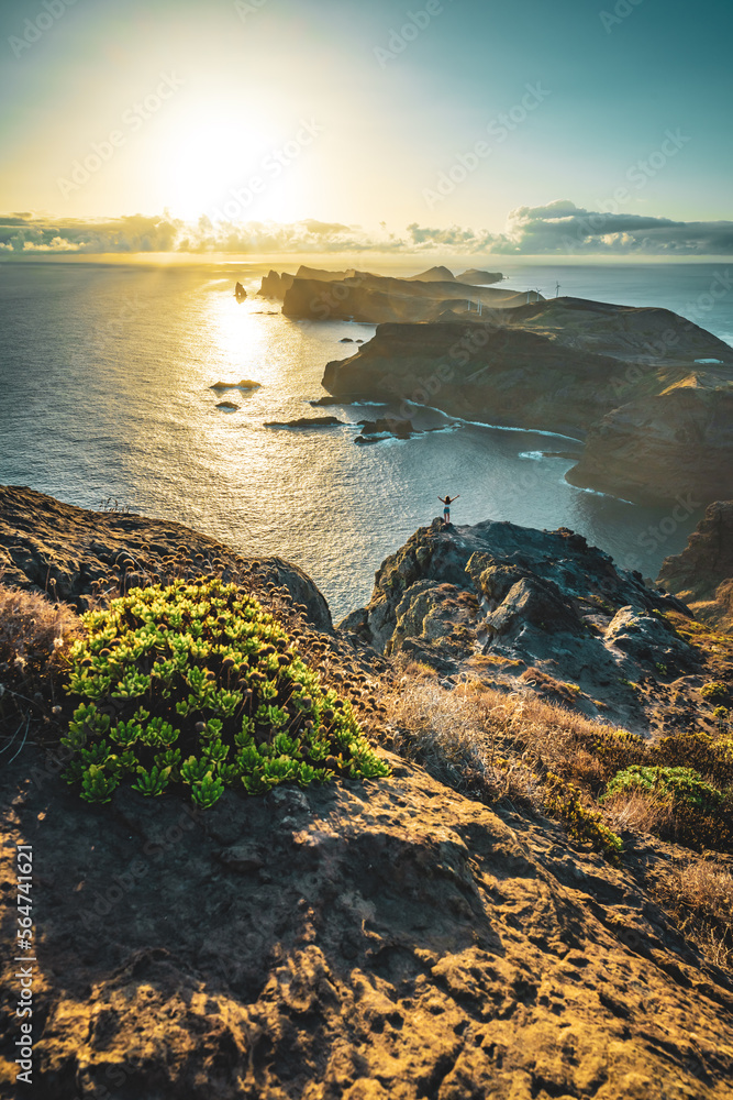 Woman enjoys panoramic view from view point on a steep cliff over the seascape and along the rugged foothills of Madeira coast at sunrise. Ponta do Bode, Madeira Island, Portugal, Europe.