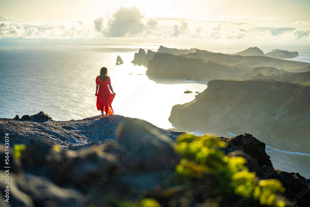 Fototapeta premium Woman in red dress enjoys panoramic view from steep cliff over seascape and along rugged foothills of Madeira coast at sunrise. Ponta do Bode, Madeira Island, Portugal, Europe.