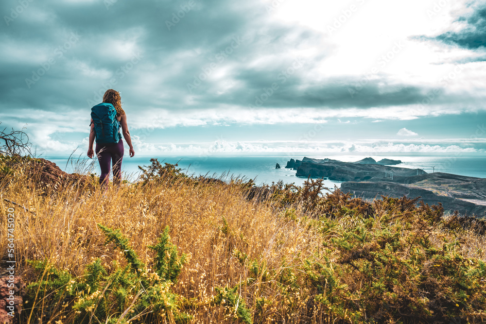 Fototapeta premium Backpacker woman enjoys hike along a steep cliff overlooking the sea and the rugged foothills of Madeira's coast in the morning. Ponta do Bode, Madeira Island, Portugal, Europe.