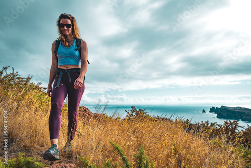 Backpacker woman enjoys hike along a steep cliff overlooking the sea and the rugged foothills of Madeira's coast in the morning. Ponta do Bode, Madeira Island, Portugal, Europe.
