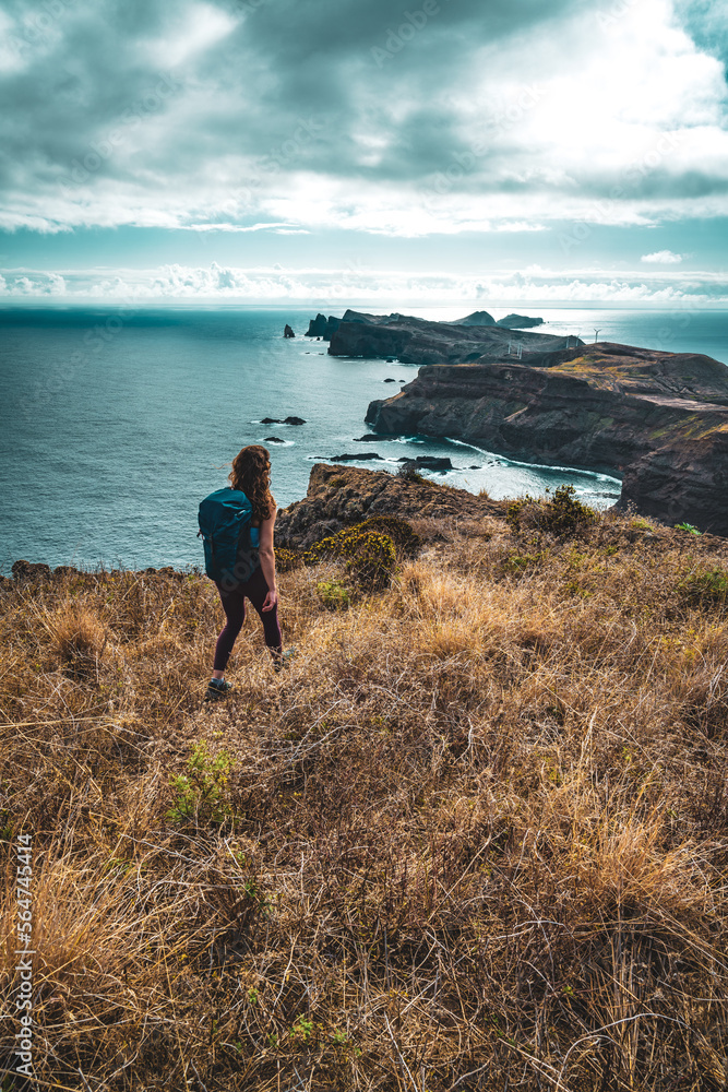 Fototapeta premium Backpacker woman enjoys hike along a steep cliff overlooking the sea and the rugged foothills of Madeira's coast in the morning. Ponta do Bode, Madeira Island, Portugal, Europe.
