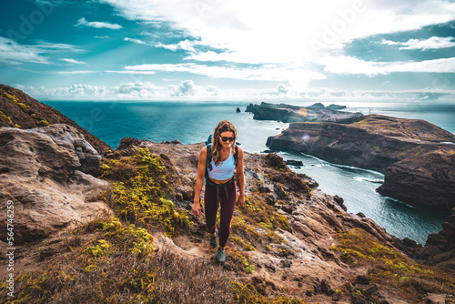 Backpacker woman enjoys hike along a steep cliff overlooking the sea and the rugged foothills of Madeira's coast in the morning. Ponta do Bode, Madeira Island, Portugal, Europe. © Michael