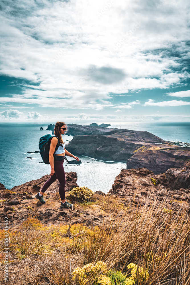 Fototapeta premium Backpacker woman enjoys hike along a steep cliff overlooking the sea and the rugged foothills of Madeira's coast in the morning. Ponta do Bode, Madeira Island, Portugal, Europe.