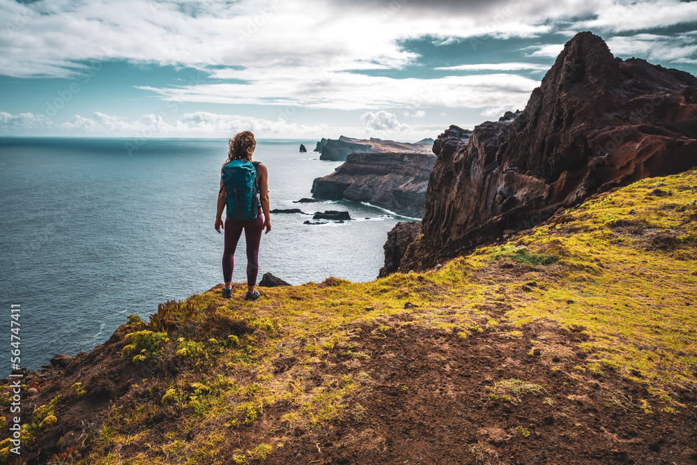 Backpacker woman enjoys panoramic view from a steep cliff overlooking the sea and the rugged foothills of Madeira's coast in the morning. Ponta do Bode, Madeira Island, Portugal, Europe.