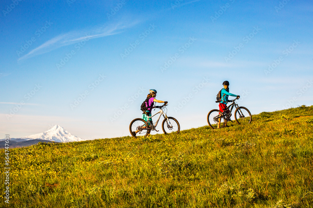 COLUMBIA RIVER GORGE, OR, USA. Two young women push mountain bikes up a single-track trail through an open meadow. COLUMBIA RIVER GORGE, OR, USA. Two young women push mountain bikes up a single-track trail through an open meadow.