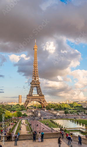 The Eiffel Tower with dark clouds and blue sky overhead glows with the summer's evening sunlight.