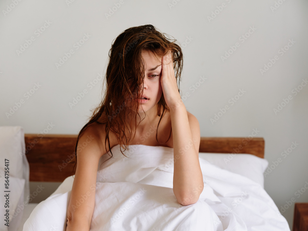 A woman sits on a bed in the bedroom and covers her body with a blanket, early awakening, sleepy appearance, rubbing her eyes from sleep, fatigue and depression