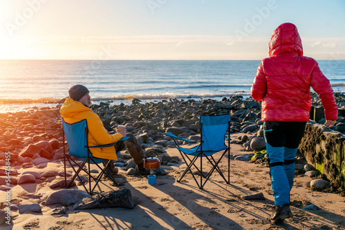 Two people on beach chairs together on winter beach, sunny, blue sky horizon. British cold winter. Local tourism concept. © Iryna