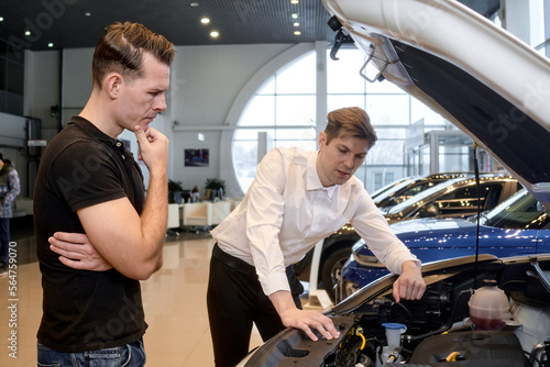 caucasian customer and salesman opened car hood and discuss all nuances concerning the car. in modern auto dealership. manager consultant showing car engine to handsome young customer, talking