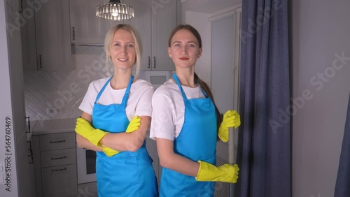 Attractive two young Caucasian white women cleaners from cleaning company in blue aprons, yellow gloves and uniforms smiling at camera in background of kitchen and quari crossing their arms. photo