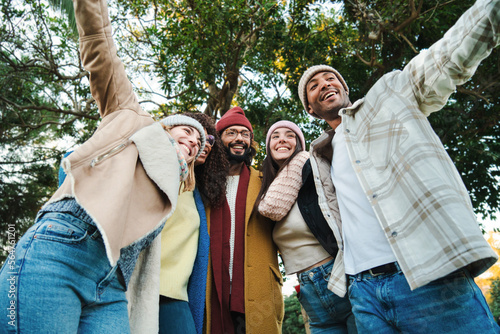 A group of multiracial young friends having fun together in a park doing autumn weekend activities wearing coats and hats. Five happy people smiling and walking outdoors. Lifestyle concept. High