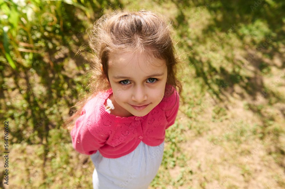 Close-up cute little girl in blue summer dress and pink bolero, smiles looking at camera, standing in nature background Close-up cute little girl in blue summer dress and pink bolero, smiles looking at camera, standing in nature background