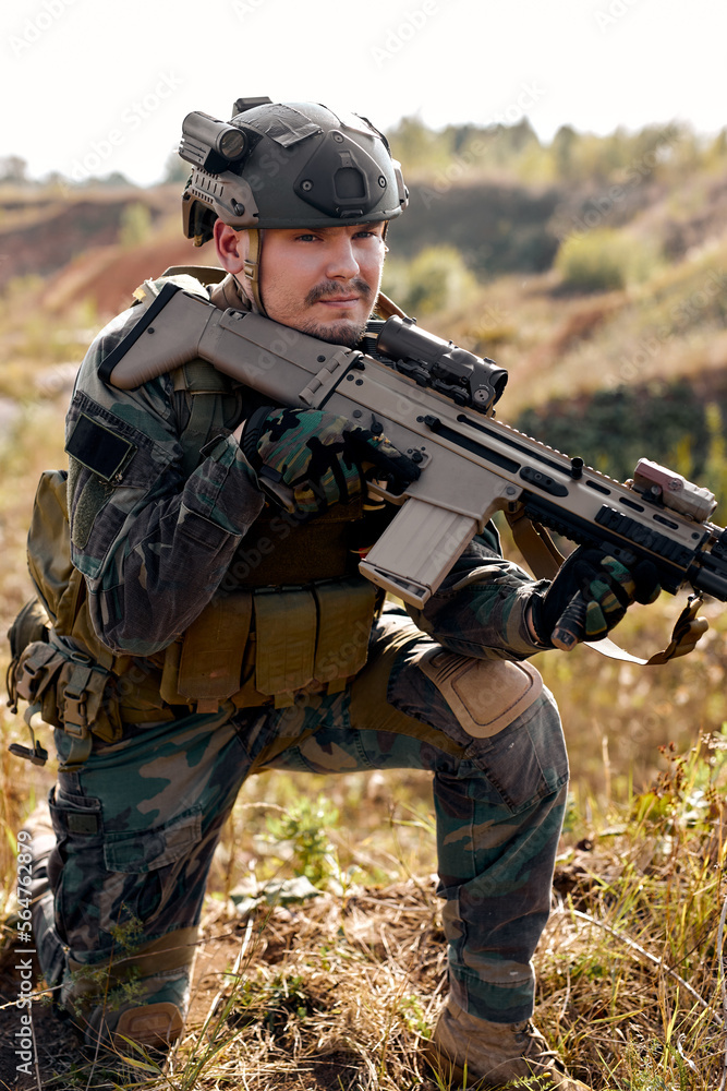 Fototapeta premium Brave caucasian male in combat uniform is standing in nature alone waiting for operation, strong soldier man in combat uniform holding rifle weapon in hands, posing, leaned on one knee