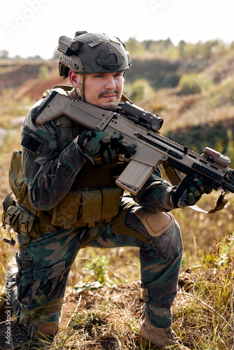 Brave caucasian male in combat uniform is standing in nature alone waiting for operation, strong soldier man in combat uniform holding rifle weapon in hands, posing, leaned on one knee
