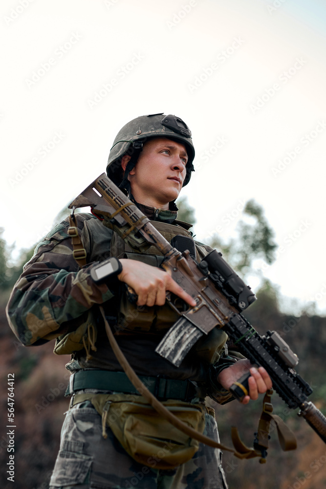 portrait of fully equipped, caucasian soldier in camouflage uniform ready to fire, holding rifle. military operation in action, standing in mountains, looking at side confidently,wait for enemy