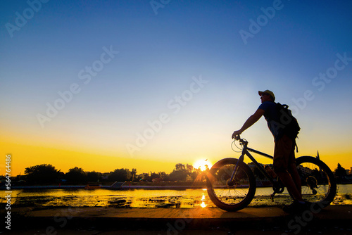 silhouette of a cyclist with a fatbike standing on the river bank at sunset