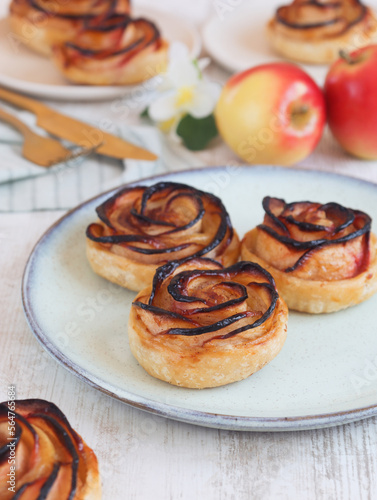 A baked mini caramelized apple rose tarts rolled in a rose form with puff pastry in a plate with knife and fork and its fresh fruit in background. 