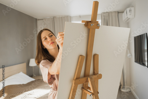 Young woman artist painting on canvas on the easel at home in bedroom - art and creativity concept