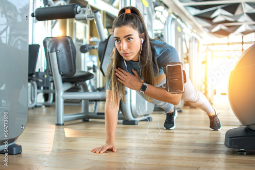 Young sportswoman doing one hand plank exercise in gym. Beautiful sporty woman with ponytail listening to music doing plank on one hand.
