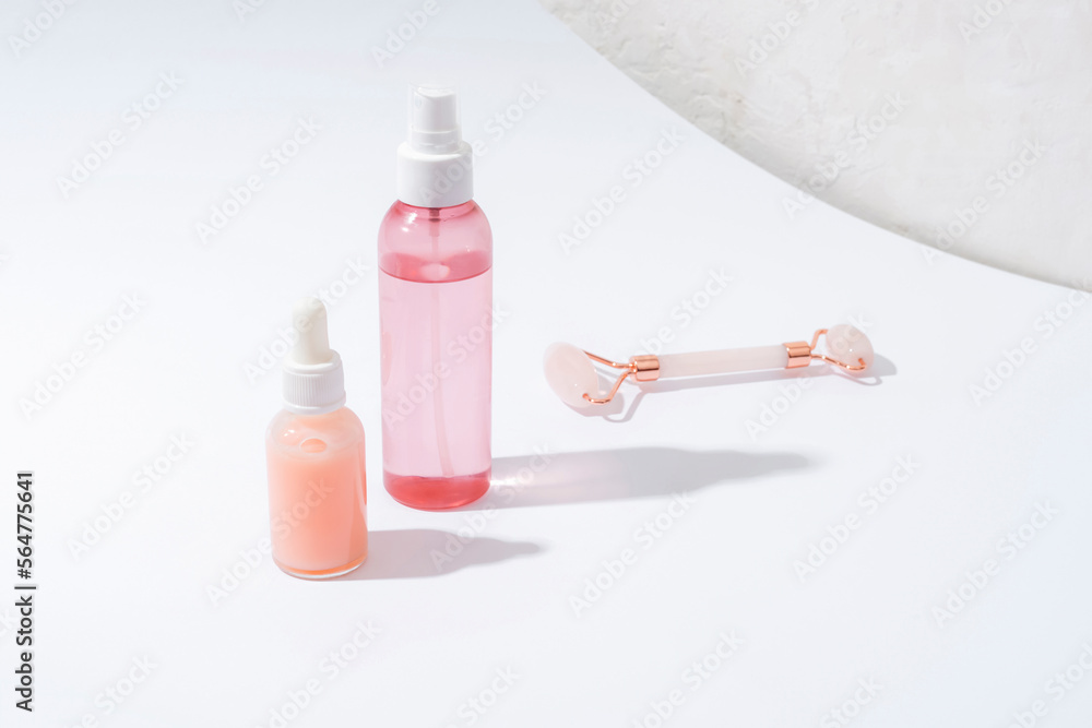 Pink cosmetic bottles and rose quartz crystal facial roller on white background with sharp shadows. Natural cosmetics, skin care concept. Front view Pink cosmetic bottles and rose quartz crystal facial roller on white background with sharp shadows. Natural cosmetics, skin care concept. Front view