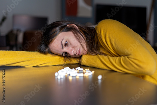 Adult melancholic female with long dark hair in yellow sweater leaning head on hand at table with pile of antidepressant pills at home photo
