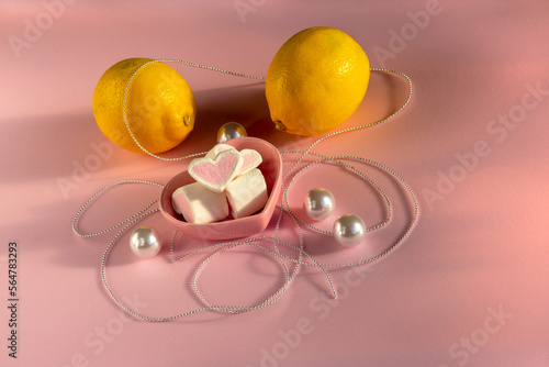 Small marshmallows in shape of heart in small pink saucer in shape of heart. On pink background. Still life in pastel colors. Decorated with lemons, large pearls