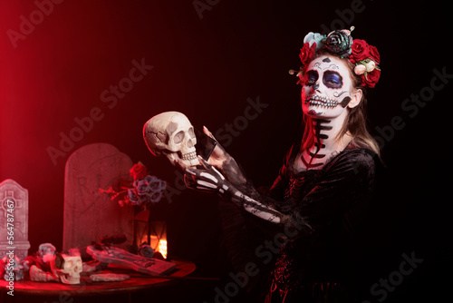 Lady of death holding smartphone for skull, acting creepy like santa muerte on mexican holiday ritual. Wearing black and white make up to celebrate dios de los muertos halloween tradition.