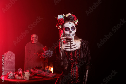 Goddess of death answering smartphone call in studio, wearing skull make up and body art to celebrate santa muerte on day of the dead. Looking like lady of dead on holy traditional ritual.