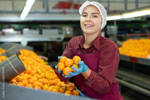 Portrait of cheerful young girl sorting ripe orange mandarins on conveyor line of factory for processing agricultural produce photo