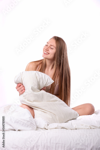 Beautiful young caucasian brown-haired girl in pink sexy silk pijama sitting on a white bed and holding a pillow and smiling on a white isolated background shot in studio