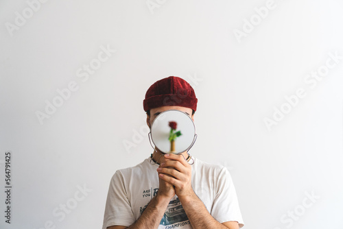 Young man wearing a red brimless hat holding a mirror in front of his face reflecting a flower with white background. Artistic photo. photo