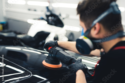 Rear view of white man in protective face mask polishing top of black car using professional buffer tool. Car detailing process. Blurred foreground. Horizontal indoor shot. High quality photo © PoppyPix