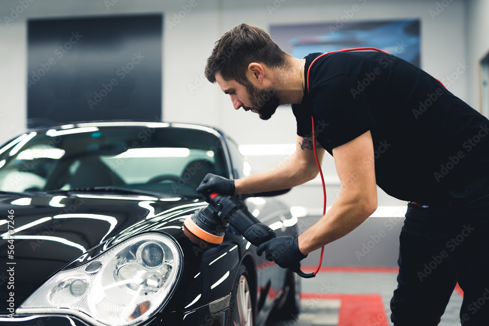 Caucasian man leaning over the front of black car using professional buffer tool to polish fresh paintwork. Car detailing process. Horizontal indoor shot. High quality photo