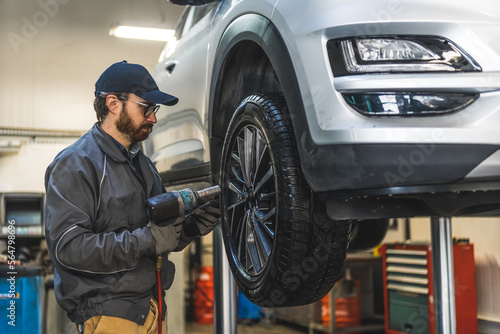 Medium shot of a mechanic standing by a car held on a lift and repairing its wheel with an impact wrench. Repair shop concept. High quality photo