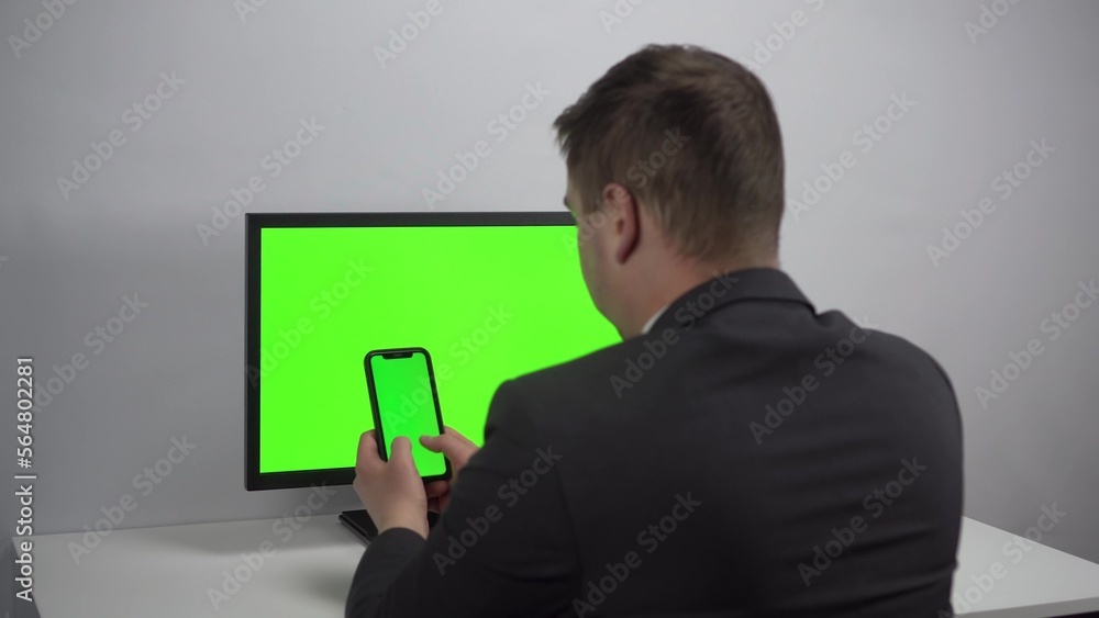 A young businessman sits at a computer and a phone in his hands with green screens. A man works in an office in a jacket. View from the back. A young businessman sits at a computer and a phone in his hands with green screens. A man works in an office in a jacket. View from the back.