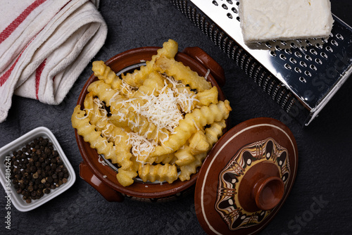 A traditional Bulgarian dish  made of potato fries topped with shredded feta cheese and served in a clay bowl called gyuveche. The picture shows a grater and chunk of feta cheese on top. photo