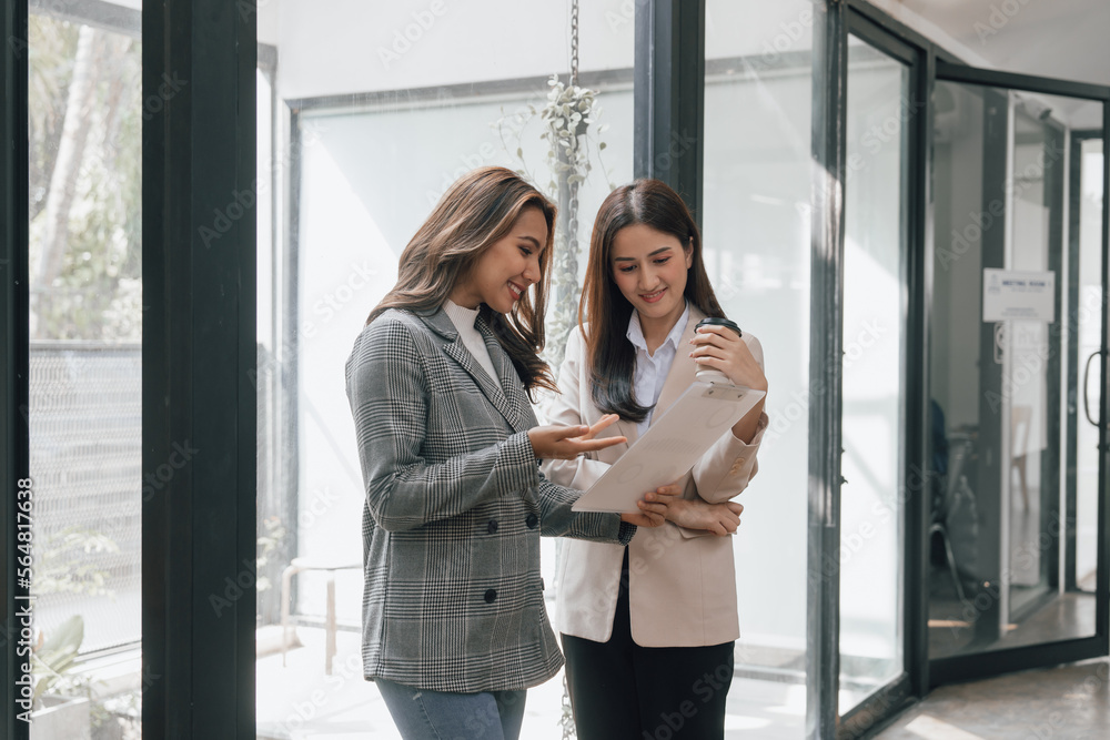 Two young pretty asia business woman in suit talking together in modern office workplace, Thai woman, southeast asian, Standing Two young pretty asia business woman in suit talking together in modern office workplace, Thai woman, southeast asian, Standing