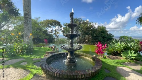 Classic water fountain in a beautiful garden with green water pond,flowers, trees, purple flowers, blue sky, on the horizon the city of Caruaru state of Pernambuco